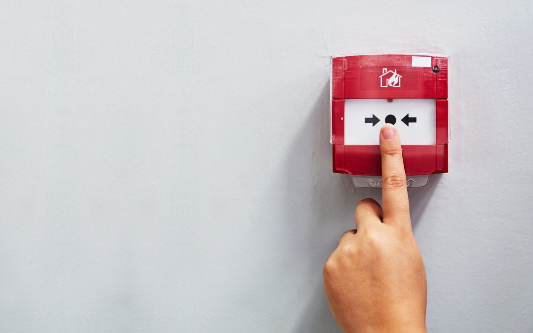 Person pressing red fire alarm call point button on white wall with finger